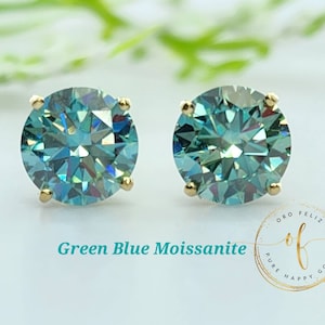 May include: A pair of round, green-blue moissanite stud earrings. The gemstones are set in gold prongs. The text "Green Blue Moissanite" is displayed in a teal font. The earrings are displayed on a white background.