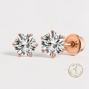 May include: A pair of rose gold stud earrings with a sparkling, round, clear gemstone. The earrings have a six-prong setting and a screw-back closure. The earrings are set against a white background.
