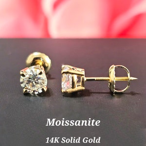 May include: A pair of gold stud earrings with round, clear moissanite stones. The earrings are made of 14K solid gold. Moissanite is a gemstone that is known for its brilliance and durability.