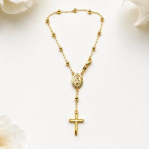 May include: Gold-toned rosary bracelet featuring a cross pendant and a central medallion. The bracelet has small, round beads and a clasp closure. The design is simple and elegant, suitable for religious occasions or as a symbol of faith.