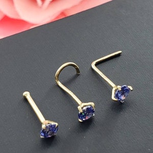 May include: Three gold nose rings with small, round, purple gemstones. The rings are on a black surface.