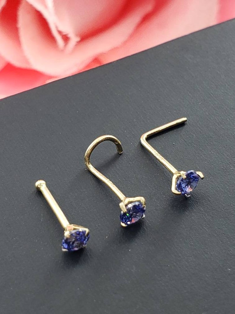 May include: Three gold nose rings with small, round, purple gemstones. The rings are on a black surface.