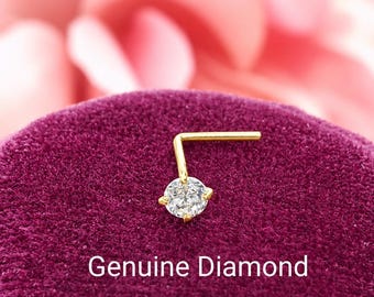 May include: An L-shaped nose stud featuring a clear, round-cut stone set in a gold-colored metal prong setting. The text Genuine Diamond is displayed below the jewelry piece, which is resting on a deep purple, textured velvet surface.