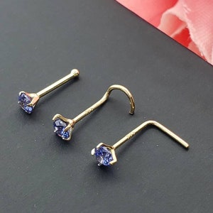 May include: Three gold nose rings with a small blue gemstone on each. The rings are different styles, one is straight, one is curved, and one is L-shaped.
