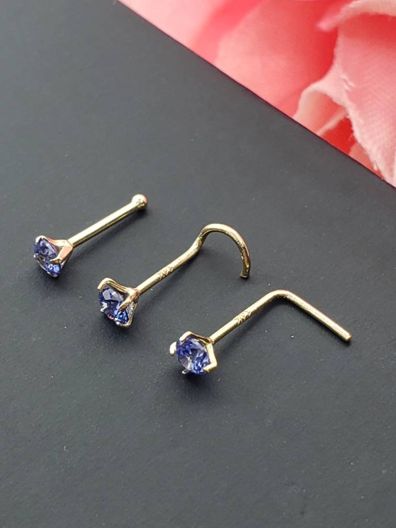 May include: Three gold nose rings with a small blue gemstone on each. The rings are different styles, one is straight, one is curved, and one is L-shaped.