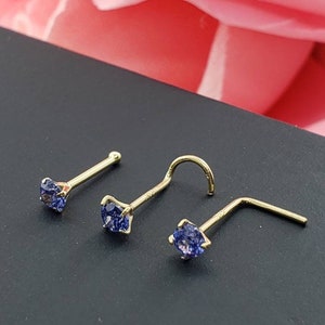 May include: Three gold nose rings with blue gemstone accents. The rings are on a dark surface.