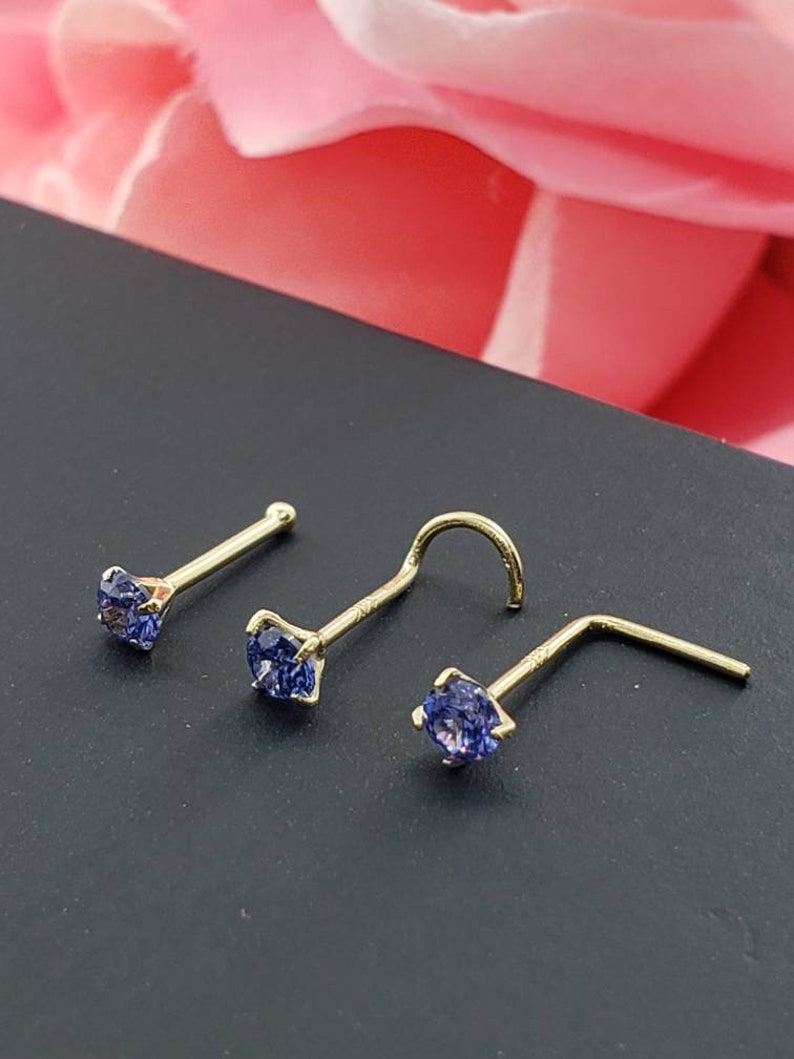 May include: Three gold nose rings with blue gemstone accents. The rings are on a dark surface.