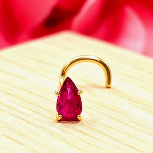 May include: A gold nose stud featuring a pear-shaped, deep pink gemstone held by four small prongs. The jewelry has a curved post design, showcasing a polished metallic finish and a vibrant, faceted stone set in a classic teardrop orientation.