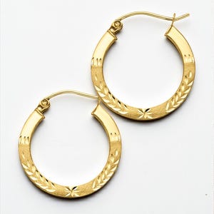 May include: A pair of gold hoop earrings. The earrings have a polished, flat surface on the top and bottom, with a textured, patterned design in the center. The pattern includes a leaf-like design and starbursts.