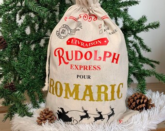 May include: A beige canvas Santa sack with red and gold lettering. The sack says "Livraison Rudolph Express Pour Romaric". There are reindeer and a sleigh at the bottom of the sack.