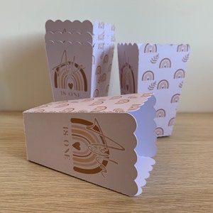 Peut inclure: Plusieurs boîtes à popcorn rose clair avec des bords festonnés. Les boîtes présentent un motif arc-en-ciel avec le nom "Ayla" et le texte "IS ONE" en or. Certaines boîtes ont un motif arc-en-ciel répété.