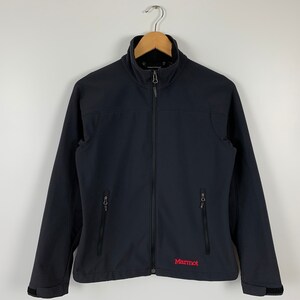Può includere: Una giacca softshell nera Marmot con chiusura a zip e due tasche con zip sul davanti. La giacca ha un colletto alto e un logo Marmot rosso sul lato inferiore sinistro.
