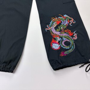 May include: Black cargo pants with a colorful embroidered dragon design on the lower leg. The dragon features red, green, and purple details. The pants have a drawstring at the ankle and a cargo pocket.