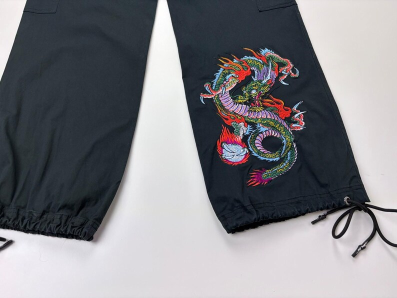 May include: Black cargo pants with a colorful embroidered dragon design on the lower leg. The dragon features red, green, and purple details. The pants have a drawstring at the ankle and a cargo pocket.