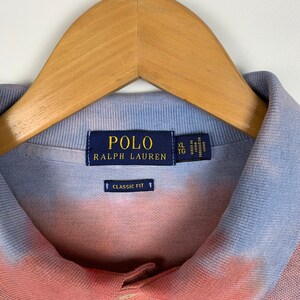 Puede incluir: Una camiseta polo de color azul y rosa tie-dye con una etiqueta de Polo Ralph Lauren. La etiqueta dice "Polo Ralph Lauren" y "Classic Fit". La etiqueta de talla dice "XL TG" y "Hecho en China".
