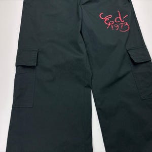 May include: Dark green cargo pants with two large pockets on the legs. The pants have a relaxed fit and feature the text "Ed 1973" in a red, stylized font on the upper right leg. The pants are made of a durable fabric.
