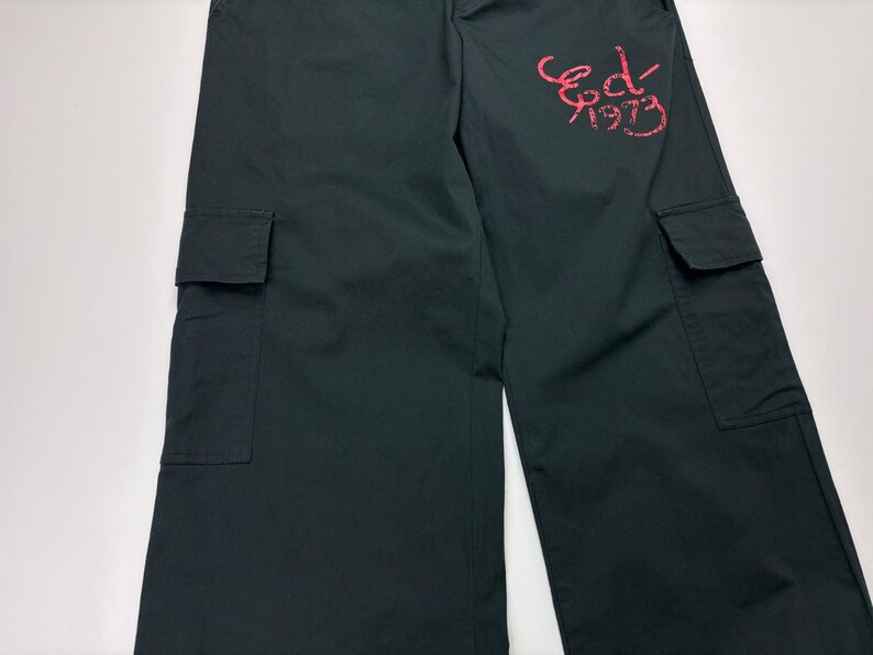 May include: Dark green cargo pants with two large pockets on the legs. The pants have a relaxed fit and feature the text "Ed 1973" in a red, stylized font on the upper right leg. The pants are made of a durable fabric.