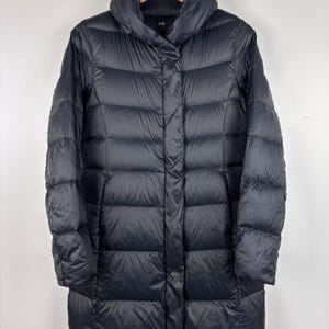 May include: A black, long puffer coat with a high collar, hanging on a wooden hanger. The coat has a quilted design and a button closure down the front. The coat is knee-length.