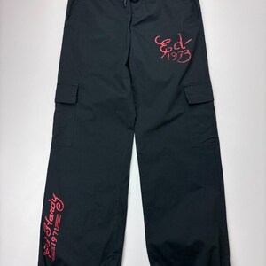 May include: Black cargo pants with a drawstring waist and ankle ties. The pants feature two cargo pockets and red embroidered text that reads "Ed Hardy 1971" and "Ed 1973".