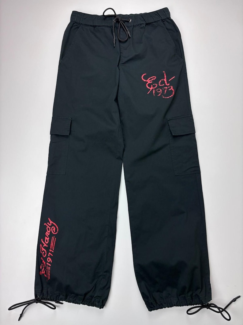 May include: Black cargo pants with a drawstring waist and ankle ties. The pants feature two cargo pockets and red embroidered text that reads "Ed Hardy 1971" and "Ed 1973".