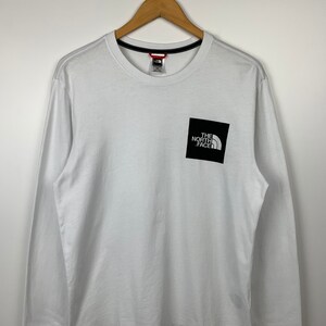 May include: White long-sleeve t-shirt with a black square logo that says "The North Face" in white lettering.