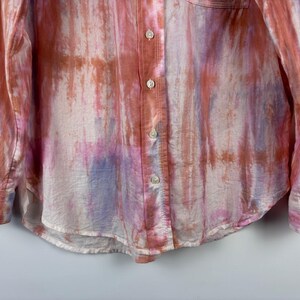 Pode incluir: Camisa de manga comprida com um padr&atilde;o tie-dye em tons de rosa, laranja e roxo. A camisa tem uma frente abotoada, uma gola cl&aacute;ssica e um bolso no peito. O tecido parece leve e ligeiramente transparente.