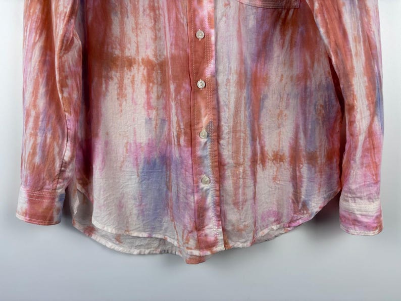 Pode incluir: Camisa de manga comprida com um padr&atilde;o tie-dye em tons de rosa, laranja e roxo. A camisa tem uma frente abotoada, uma gola cl&aacute;ssica e um bolso no peito. O tecido parece leve e ligeiramente transparente.