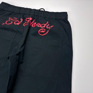 May include: Black pants with two back pockets and an elastic waistband. The red "Ed Hardy" logo is printed across the back. The pants are made of a durable fabric and have a drawstring closure.