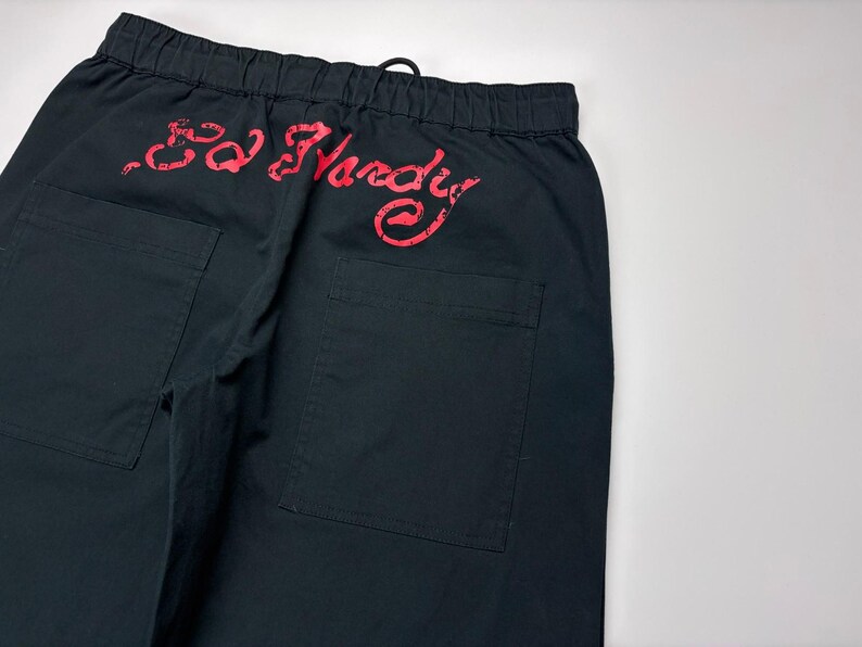 May include: Black pants with two back pockets and an elastic waistband. The red "Ed Hardy" logo is printed across the back. The pants are made of a durable fabric and have a drawstring closure.