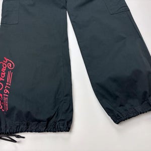 May include: Black cargo pants with a drawstring hem and pockets. The pants feature the text "Ed Hardy 1971" in red script on the left leg. The pants are made of a lightweight material.
