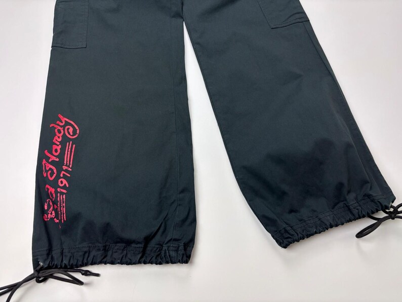 May include: Black cargo pants with a drawstring hem and pockets. The pants feature the text "Ed Hardy 1971" in red script on the left leg. The pants are made of a lightweight material.