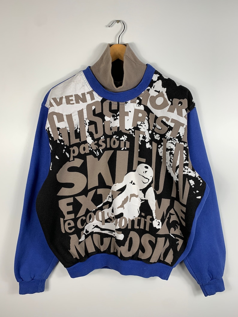 Puede incluir: Una sudadera azul, negra y blanca con un estampado gr&aacute;fico de un esquiador y las palabras "Glisse", "Passion", "Ski Fun", "Extreme", "Le coq sportif" y "Microski".