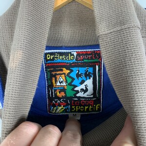 Puede incluir: Una camisa azul y marr&oacute;n con cuello y una etiqueta colorida que dice "Dr&ocirc;les de sport" y "La coq Sportif" con un logotipo de gallo. La etiqueta tambi&eacute;n tiene la talla "M" impresa.