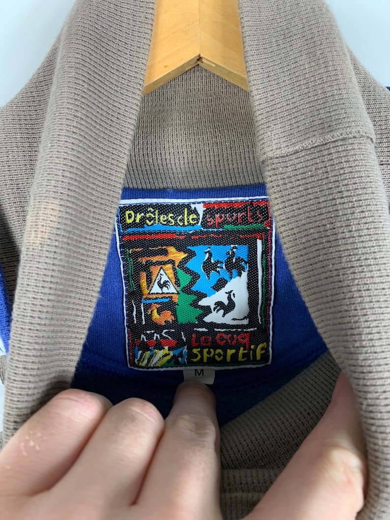 Puede incluir: Una camisa azul y marr&oacute;n con cuello y una etiqueta colorida que dice "Dr&ocirc;les de sport" y "La coq Sportif" con un logotipo de gallo. La etiqueta tambi&eacute;n tiene la talla "M" impresa.