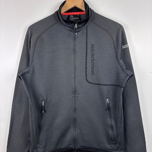 May include: A dark grey zip-up jacket with the text "Peak Performance" down the front. The jacket features a black collar with a red accent, two zipped pockets, and the letters "R&D" on the sleeve.