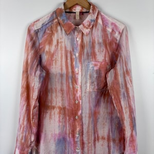 Pode incluir: Camisa abotoada de manga comprida com um padr&atilde;o tipo aquarela em tons de rosa, laranja e roxo. A camisa, com bolso no peito, est&aacute; pendurada em um cabide de madeira.