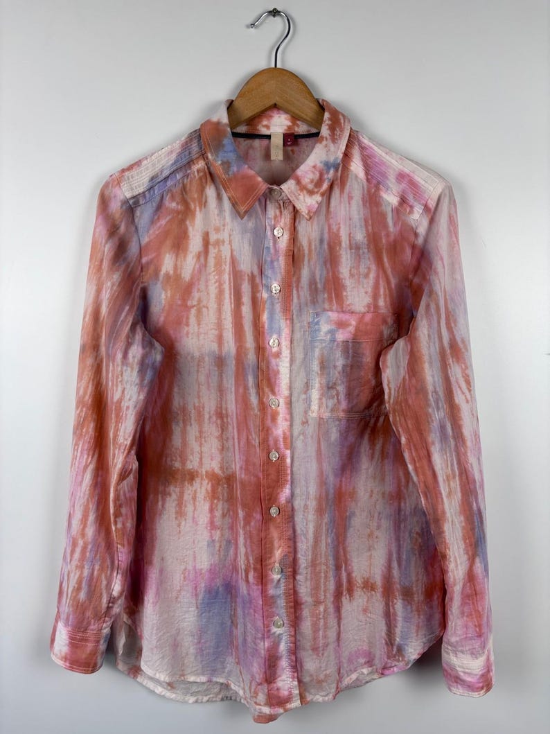 Pode incluir: Camisa abotoada de manga comprida com um padr&atilde;o tipo aquarela em tons de rosa, laranja e roxo. A camisa, com bolso no peito, est&aacute; pendurada em um cabide de madeira.