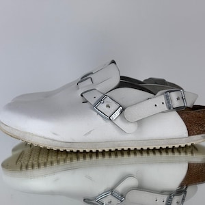 May include: White leather clogs with adjustable straps and silver buckles. The clogs have a cork and rubber sole with a brown accent on the heel. The shoes are reflected in a shiny surface.