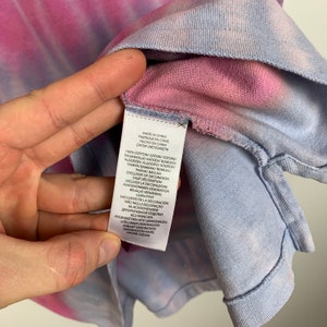 Puede incluir: Una camisa de color rosa y azul con estampado tie-dye con una etiqueta blanca que dice "Hecho en China" y enumera los materiales y las instrucciones de cuidado en varios idiomas.