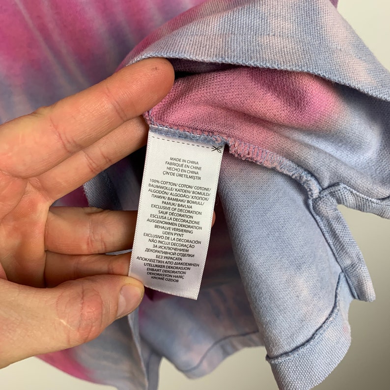 Puede incluir: Una camisa de color rosa y azul con estampado tie-dye con una etiqueta blanca que dice "Hecho en China" y enumera los materiales y las instrucciones de cuidado en varios idiomas.