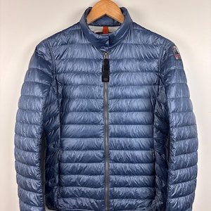 Puede incluir: Chaqueta acolchada azul marino con cuello alto y cremallera completa. La chaqueta tiene un ligero brillo y un pequeño parche con el logotipo en la manga superior izquierda. Cuelga de una percha de madera sobre un fondo blanco.