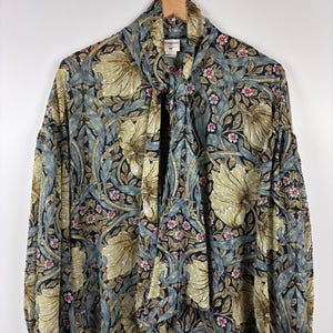 May include: A patterned blouse in olive green, blue, and black, hanging on a wooden hanger. The blouse has a tie-neck and gathered sleeves. The floral design features large flowers and small pink blossoms. The fabric appears to be lightweight.