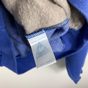 Puede incluir: Una etiqueta blanca con costuras azules y el texto "fabriqu&eacute; en FRANCE" y "le coq sportif PARIS FRANCE" unida a una sudadera azul.