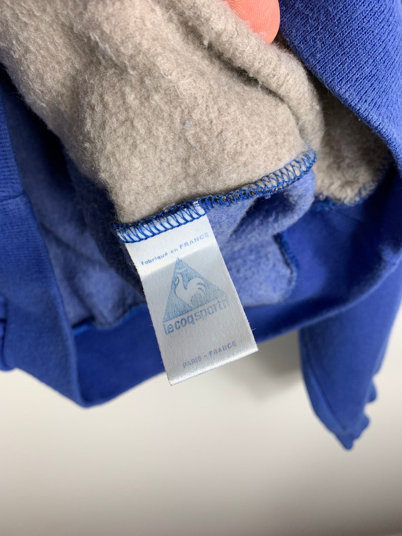 Puede incluir: Una etiqueta blanca con costuras azules y el texto "fabriqu&eacute; en FRANCE" y "le coq sportif PARIS FRANCE" unida a una sudadera azul.