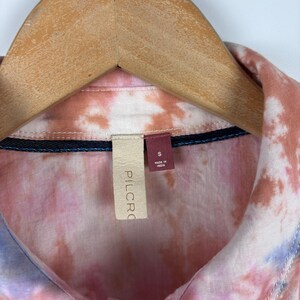 Pode incluir: Uma camisa tie-dye em tons de rosa, p&ecirc;ssego e azul, pendurada em um cabide de madeira. A camisa tem gola e uma etiqueta com "PILCRO" e "S Made in India".