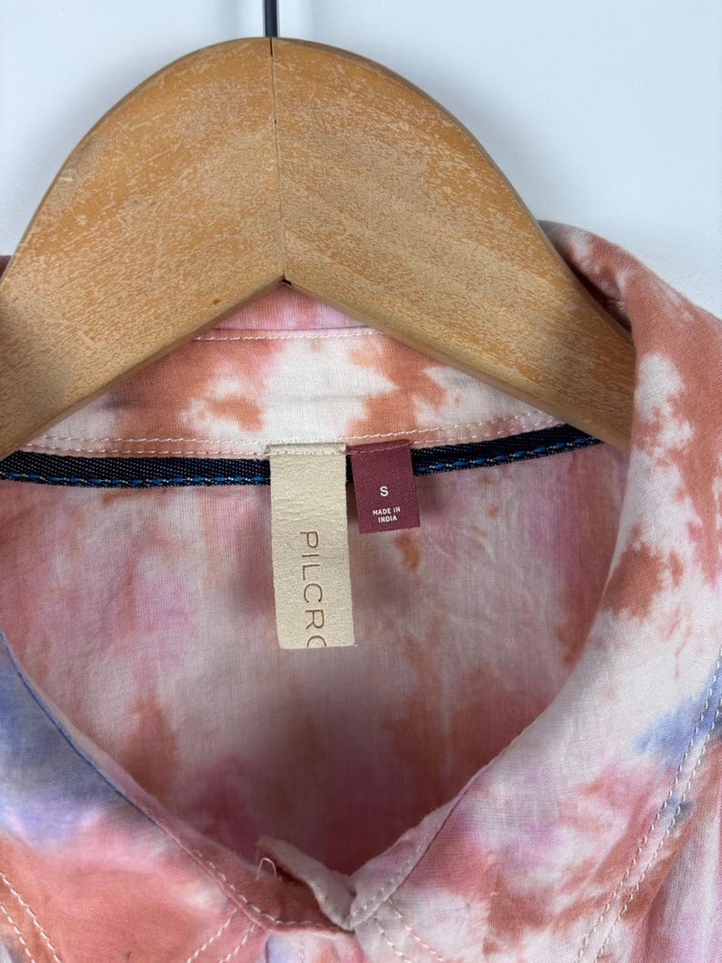 Pode incluir: Uma camisa tie-dye em tons de rosa, p&ecirc;ssego e azul, pendurada em um cabide de madeira. A camisa tem gola e uma etiqueta com "PILCRO" e "S Made in India".