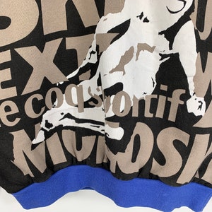 Puede incluir: Sudadera negra con un estampado gr&aacute;fico marr&oacute;n y blanco de una persona haciendo una parada de manos. El texto "le coq sportif" est&aacute; impreso en marr&oacute;n en la sudadera. La sudadera tiene un pu&ntilde;o de canal&eacute; azul.