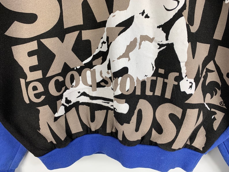 Puede incluir: Sudadera negra con un estampado gr&aacute;fico marr&oacute;n y blanco de una persona haciendo una parada de manos. El texto "le coq sportif" est&aacute; impreso en marr&oacute;n en la sudadera. La sudadera tiene un pu&ntilde;o de canal&eacute; azul.