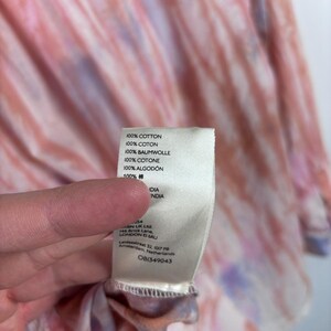 Pode incluir: Um close-up de uma pe&ccedil;a de roupa com padr&atilde;o tie-dye em tons de rosa, laranja e roxo. Uma etiqueta branca com texto em v&aacute;rios idiomas, incluindo "100% COTTON" e "OB1349043", &eacute; vis&iacute;vel.