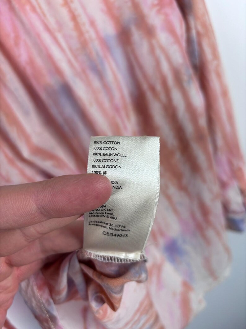 Pode incluir: Um close-up de uma pe&ccedil;a de roupa com padr&atilde;o tie-dye em tons de rosa, laranja e roxo. Uma etiqueta branca com texto em v&aacute;rios idiomas, incluindo "100% COTTON" e "OB1349043", &eacute; vis&iacute;vel.
